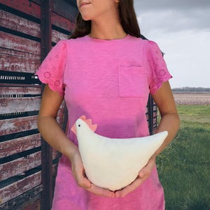 Może przedstawiać: Biały pluszowy kurczak z czerwonym i białym grzebieniem trzymany jest przed osobą ubraną w różową koszulkę z kieszenią. Koszula ma rękawy z ażurowej koronki. Tło obejmuje zwietrzałą drewnianą konstrukcję i pole.