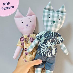 以下が含まれることがあります： 刺繍された顔の特徴を持つ2体の手作り猫人形。左の人形は薄紫色で花柄のドレスを着ています。右の人形は緑と白のギンガムチェック柄で、白い葉のプリントが施されたダークブルーのシャツとダークブルーのショートパンツを着用しています。