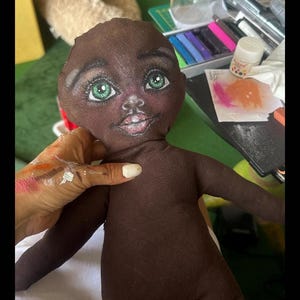 May include: A brown fabric doll with a painted face. The doll has green eyes, a pink mouth, and a brown body. The doll is sitting on a white surface.