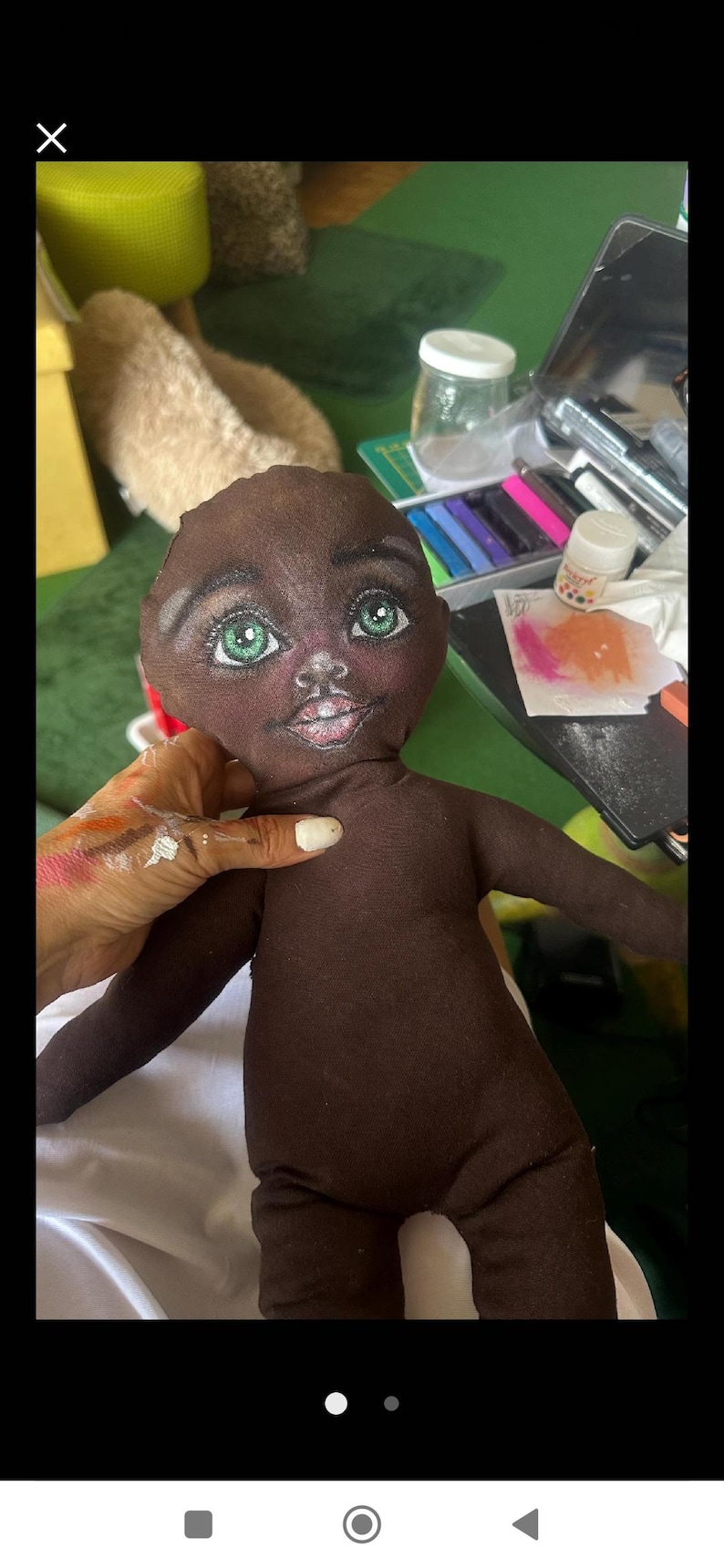May include: A brown fabric doll with a painted face. The doll has green eyes, a pink mouth, and a brown body. The doll is sitting on a white surface.