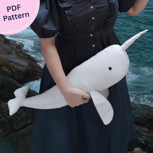 May include: A white plush narwhal toy with a single horn, a small black eye, and a tail fin. The toy is held against a dark dress. A circular pink badge in the top left corner contains the white text PDF Pattern.