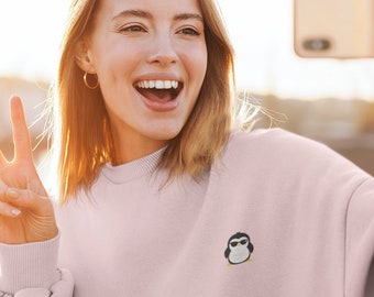 May include: A woman wearing a pink sweatshirt with a small embroidered penguin design is taking a selfie with a gold phone. She is smiling and giving a peace sign with her left hand.