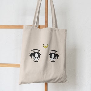 May include: A beige tote bag featuring a black and yellow anime-style eye design. The eyes are large with detailed eyelashes and a yellow crescent moon above them. The bag hangs on a wooden ladder against a white wall.