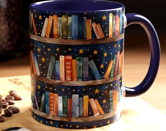 May include: A ceramic mug with a dark blue interior and handle. The exterior features a whimsical design of bookshelves filled with colorful books against a starry night sky. A spoon and a macaron are on a wooden surface.