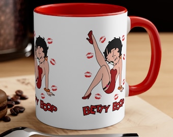 May include: White ceramic mug with a red rim and handle. The mug features a cartoon image of Betty Boop in two poses, with red lips scattered around the image. The text "Betty Boop" is printed on the mug.