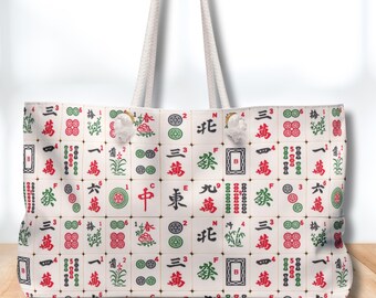 May include: A white tote bag with a repeating pattern of Mahjong tiles in red, green, and black. The bag has white rope handles and is sitting on a wooden surface.