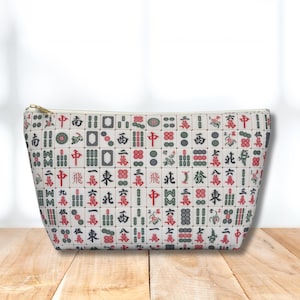 May include: A white pouch with a gold zip, patterned with red, green, and black Mahjong tiles. The pouch is displayed on a wooden surface, with a soft background.