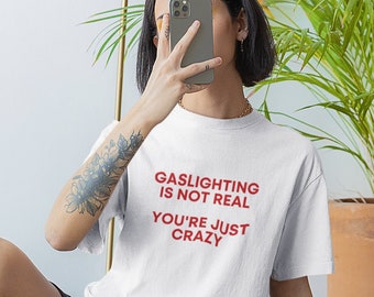 May include: White t-shirt with red text that reads "Gaslighting is not real you're just crazy". A person is wearing the t-shirt and sitting on a wooden floor.