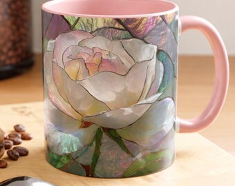 May include: A ceramic coffee mug with a pink handle and interior. The mug features a detailed floral design of a white rose with green leaves. The mug is on a wooden surface with a spoon and a macaron.