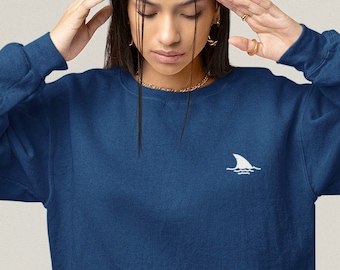 May include: A woman wearing a dark blue sweatshirt with a white shark fin logo on the left chest. She is wearing blue jeans and is standing in front of a red background.