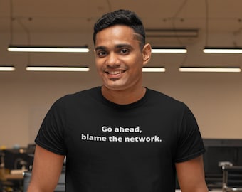 May include: A man wearing a black t-shirt with the text "Go ahead, blame the network." printed in white. He is standing in an office setting with desks and chairs in the background.