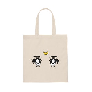 May include: A beige tote bag featuring a cartoon illustration of large anime-style eyes with long eyelashes and a yellow crescent moon above them. The eyes are black and white, with a tear drop detail.