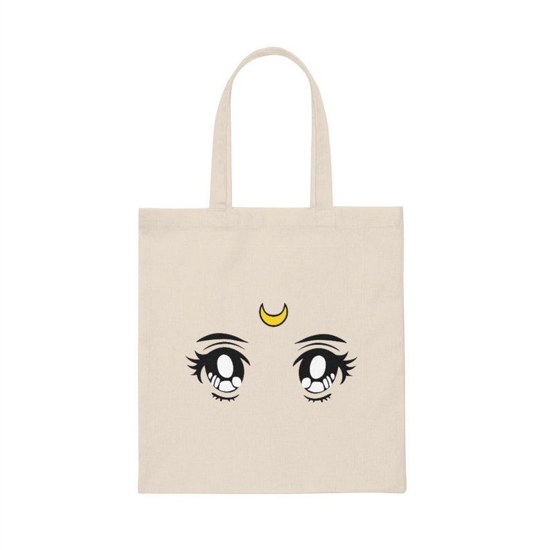 May include: A beige tote bag featuring a cartoon illustration of large anime-style eyes with long eyelashes and a yellow crescent moon above them. The eyes are black and white, with a tear drop detail.