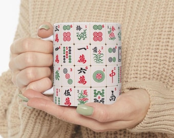 May include: White ceramic mug featuring a repeating pattern of Mahjong tiles in red, green, and black. The mug is held by a person wearing a beige sweater. The tiles display various symbols and numbers.
