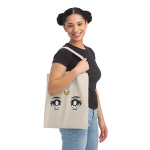 May include: A beige tote bag featuring a cartoon eye design with a yellow crescent moon. The eyes have long eyelashes and are outlined in black. The bag is held by a person wearing a black shirt and blue jeans.