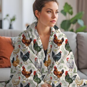 May include: A white throw blanket with a repeating pattern of watercolor chickens and green foliage. The blanket is draped over a couch and a woman is holding the blanket around her shoulders.