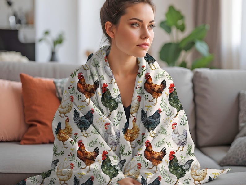 May include: A white throw blanket with a repeating pattern of watercolor chickens and green foliage. The blanket is draped over a couch and a woman is holding the blanket around her shoulders.