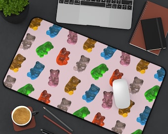 May include: A pink mouse pad with a repeating pattern of colorful gummy bear shapes. The mouse pad is on a black surface with a laptop, a notebook, and a cup of coffee.