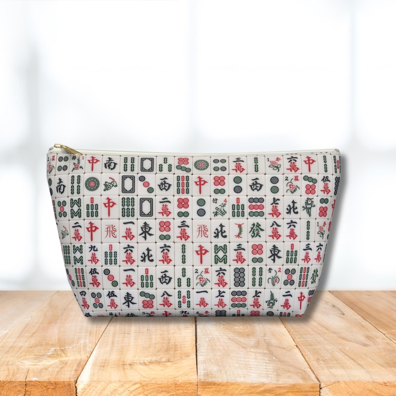 May include: A white pouch with a gold zip, patterned with red, green, and black Mahjong tiles. The pouch is displayed on a wooden surface, with a soft background.