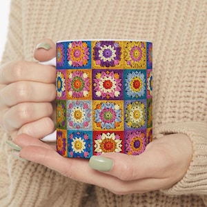 Op de afbeelding: Een witte keramische mok met een kleurrijk gehaakt patroon. Het patroon bestaat uit vierkanten in verschillende kleuren, waaronder roze, paars, geel, groen en blauw.