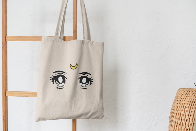 May include: A beige tote bag featuring a black and yellow anime-style eye design. The eyes are large with detailed eyelashes and a yellow crescent moon above them. The bag hangs on a wooden ladder against a white wall.