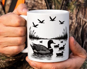 May include: White ceramic mug featuring a black and white illustration of ducks swimming in water, with flying birds and reeds. The mug is held by a person wearing a camouflage jacket.