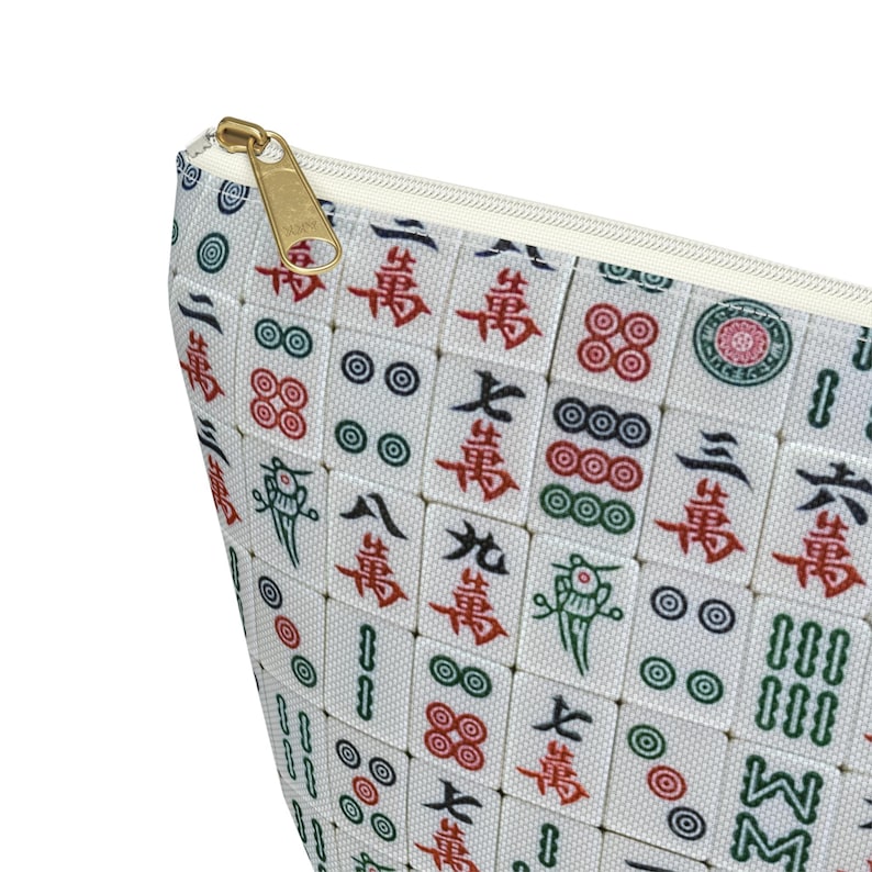 May include: A white zippered pouch with a repeating pattern of mahjong tiles. The tiles feature red, green, and black symbols and numbers. The pouch has a gold zipper.