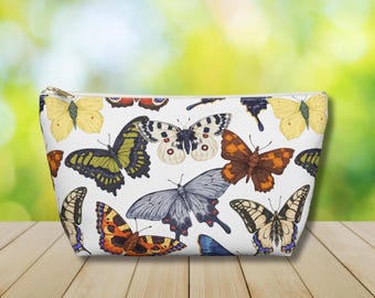 May include: A white cosmetic bag with a gold zipper, adorned with a colorful butterfly pattern. The butterflies are in various colors, including yellow, green, orange, and gray. The bag is sitting on a wooden surface.