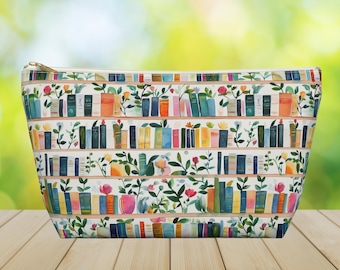 May include: A white cosmetic bag with a watercolor design of bookshelves filled with colorful books and flowers. The bag has a gold zipper and is set on a wooden surface.