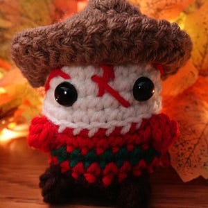 May include: A handmade crochet figure resembling a character with a brown hat, white face with red accents, black eyes, and a red and green striped sweater. The figure is standing on a wooden surface with autumn leaves in the background.