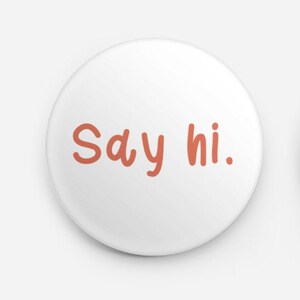 Può includere: Una spilla rotonda con sfondo bianco e il testo "Say hi." scritto in un carattere arrotondato color salmone. Il retro della spilla è in metallo argentato e presenta un meccanismo a spilla orizzontale per l'applicazione sui vestiti.