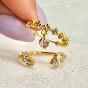 May include: Two gold rings with colorful stones. One ring has a heart-shaped charm with a pink stone. The other ring has two clusters of clear stones.