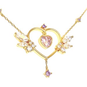 May include: A gold necklace with a heart-shaped pendant adorned with clear and purple crystals. The pendant features a smaller pink heart in the center.