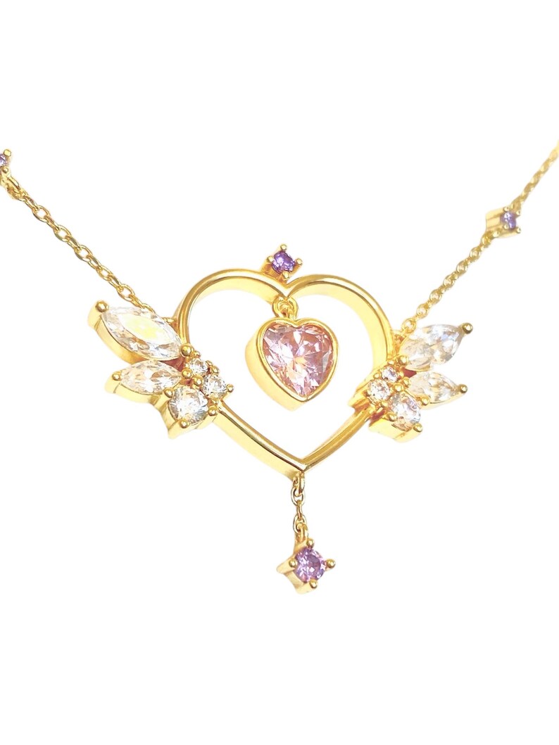 May include: A gold necklace with a heart-shaped pendant adorned with clear and purple crystals. The pendant features a smaller pink heart in the center.