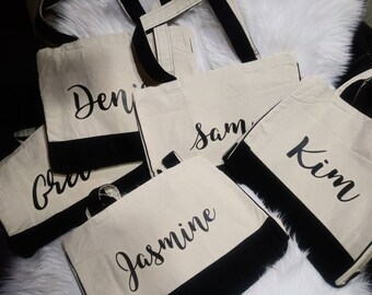 Könnte beinhalten: Fünf weiße Canvas-Tragetaschen mit schwarzen Griffen und schwarzem Besatz. Jede Tasche trägt einen Namen in schwarzer Schrift:  "Deni", "Sam", "Kim", "Gre" und "Jasmine".