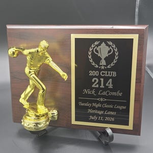 May include: A rectangular wooden plaque featuring a gold-colored bowling figure on the left and a black plate on the right. The plate displays a trophy icon, the text 200 CLUB, 214, Nick LaCombe, Tuesday Night Classic League, Heritage Lanes, July 11, 2026.