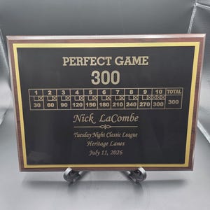 May include: A rectangular wooden plaque with a black center panel and gold border. The text reads PERFECT GAME 300, featuring a bowling scorecard, the name Nick LaCombe, Tuesday Night Classic League, Heritage Lanes, and the date July 11, 2026.