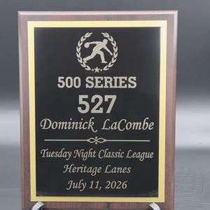 May include: A rectangular wooden plaque with a dark brown finish and a black center plate featuring gold trim. The text reads 500 SERIES, 527, Dominick LaCombe, Tuesday Night Classic League, Heritage Lanes, July 11, 2026, with a bowling graphic.