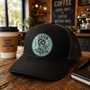 Peut inclure: Casquette de camionneur noire avec un dos en filet et un écusson circulaire représentant une femme portant des lunettes de soleil et les mots "JAVA JUNKIE". Une tasse de café avec le texte "COFFEE FUELS EVERYTHING" est à gauche.