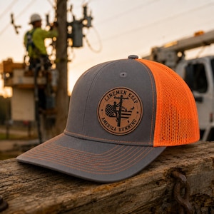 May include: A gray trucker hat with an orange mesh back and a curved brim. The hat features a leather patch with the text "Linemen Keep America Running" and an image of a lineman and the American flag.
