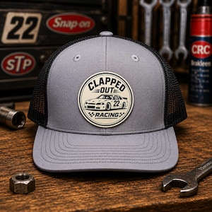 Puede incluir: Gorra trucker gris con parte trasera de malla negra y un parche circular. El parche presenta un coche de carreras y las palabras "CLAPPED OUT RACING 22". La gorra está sobre una superficie de madera.