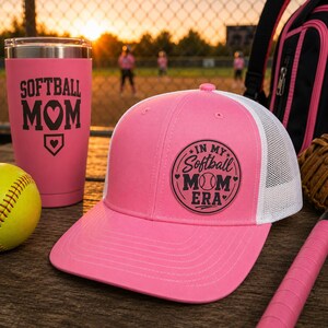 Könnte beinhalten: Eine rosa Baseballkappe mit weißem Mesh-Rücken und dem Text "IN MY Softball MOM ERA". Ein rosa Becher mit der Aufschrift "SOFTBALL MOM" und ein gelber Softball sind ebenfalls zu sehen.