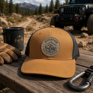 May include: A tan and black trucker hat with a circular patch that reads "Adventure Has No Pavement." The hat is on a wooden surface with a black mug, gloves, and a carabiner. A green Jeep is in the background.