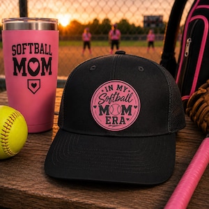 Könnte beinhalten: Schwarze Trucker-Mütze mit einem rosa runden Aufnäher mit der Aufschrift "IN MY Softball MOM ERA". Ein rosa Becher mit der Aufschrift "SOFTBALL MOM" steht neben der Mütze. Ein gelber Softball und ein rosa Schläger sind ebenfalls zu sehen.