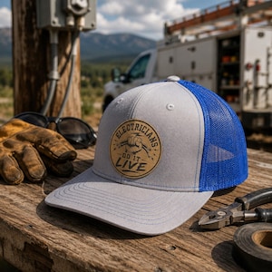 May include: A gray and blue trucker hat with the text "Electricians Do It Live" on a brown patch. The hat is on a wooden surface with work gloves, sunglasses, and tools in the background. The hat has a blue mesh back.