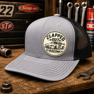 Puede incluir: Gorra de camionero gris con la parte trasera de malla negra. La gorra presenta un parche circular con las palabras "CLAPPED OUT RACING" alrededor de un gráfico de un coche de carreras y el número 22. La gorra está sobre una superficie de madera.