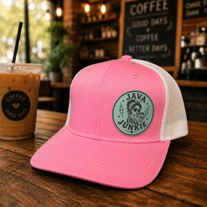 Peut inclure: Casquette rose avec un dos en filet blanc et une visière incurvée. Le chapeau porte un écusson circulaire avec le texte "JAVA JUNKIE" et une illustration d'une femme. Un verre de café glacé est visible en arrière-plan.