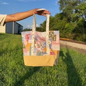May include: A patchwork tote bag featuring a variety of floral, gingham, and polka dot fabric squares in shades of pink, blue, yellow, and green. The bag has a solid tan base, two patterned handles, and a small front pocket with a rectangular label.