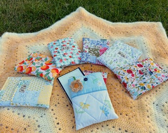 May include: A collection of colorful, quilted book sleeves and covers arranged on a crocheted blanket. The sleeves feature various floral, fruit, and patchwork patterns. A book is nestled in a light blue sleeve with a crocheted flower.