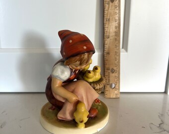 Puede incluir: Una figurita de porcelana de una niña con un vestido rojo y blanco de lunares y un sombrero rojo y blanco de lunares que sostiene una cesta de pollitos amarillos.